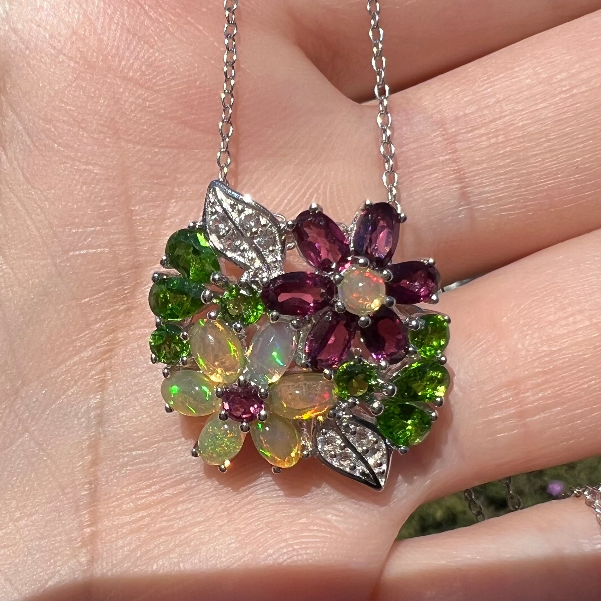 A sterling silver opal and garnet flower necklace shown close-up in the palm of a hand in sunlight.