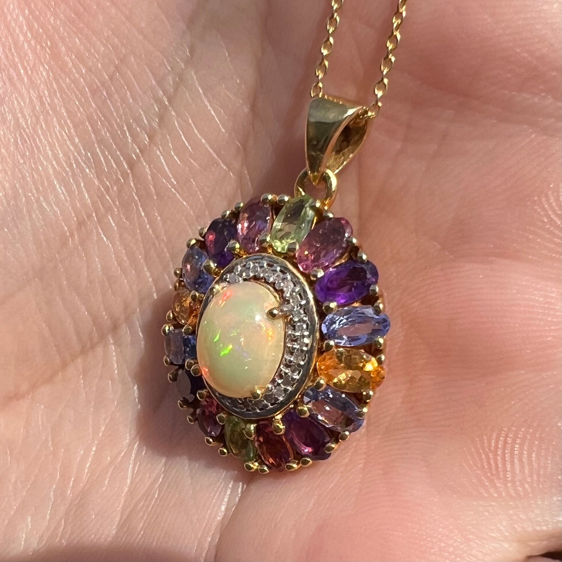 A gold plated Ethiopian fire opal and multi-gemstone halo pendant shown from an angle in sunlight.