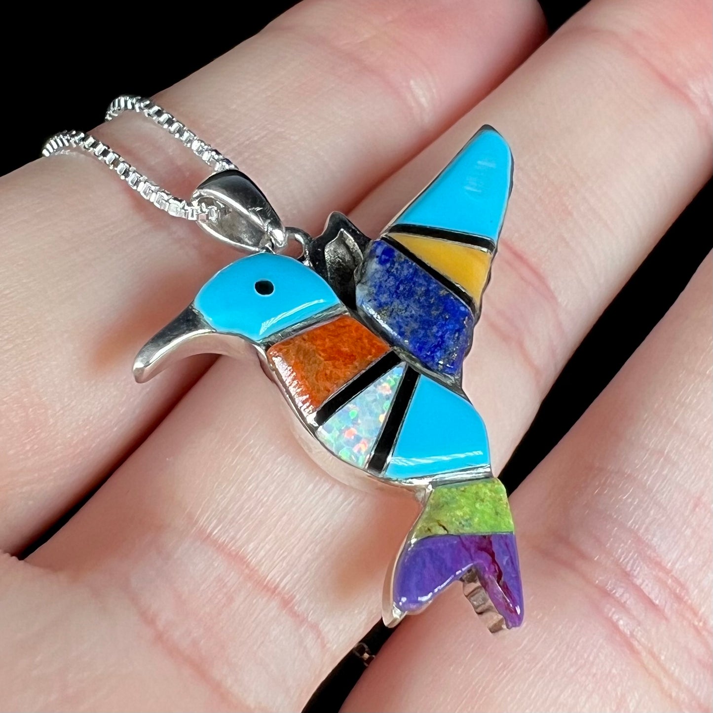A multi-stone inlay hummingbird necklace in sterling silver, shown close-up at an angle in a hand.