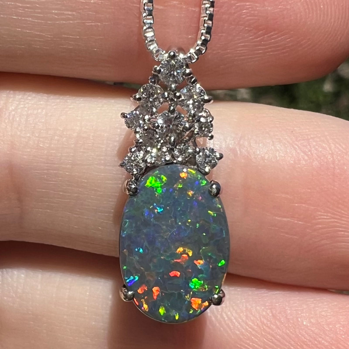 Close-up of a 2.65ct Australian black opal pendant with crown of diamond accents, shown in hand in sunlight.  Multicolor play-of-color speckles across the opal's surface.