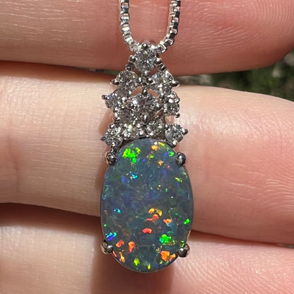 Close-up of a 2.65ct Australian black opal pendant with crown of diamond accents, shown in hand in sunlight.  Multicolor play-of-color speckles across the opal's surface.