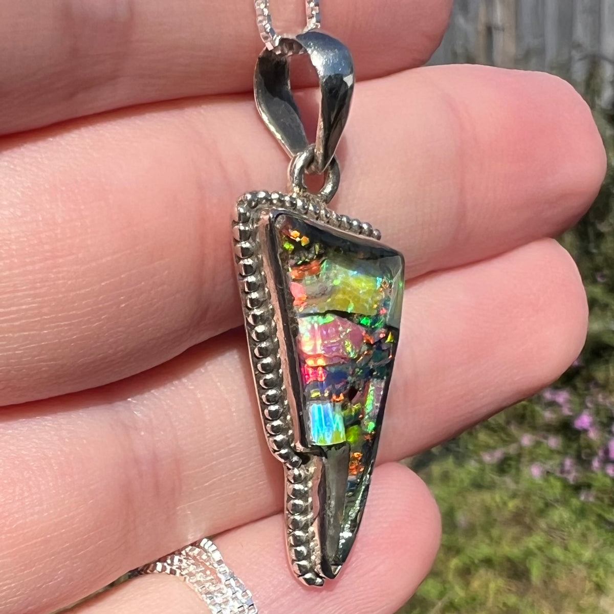 Side view of multicolor synthetic opal mosaic pendant held in sunlight, its sterling silver beaded design in focus.