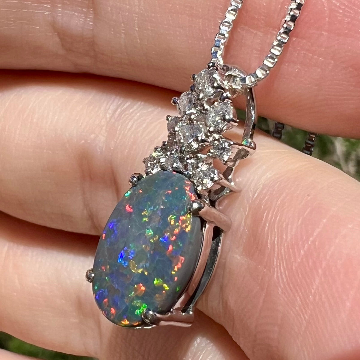 Side angle close-up of N3 black opal pendant with multicolor red and blue flashes, held in sunlight.