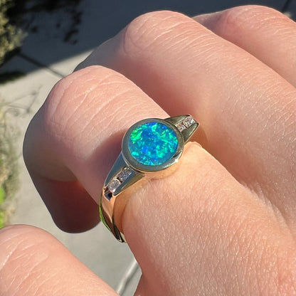 A N4 black boulder opal ring with diamond accents in yellow gold, shown close-up on a finger from an angle in sunlight.
