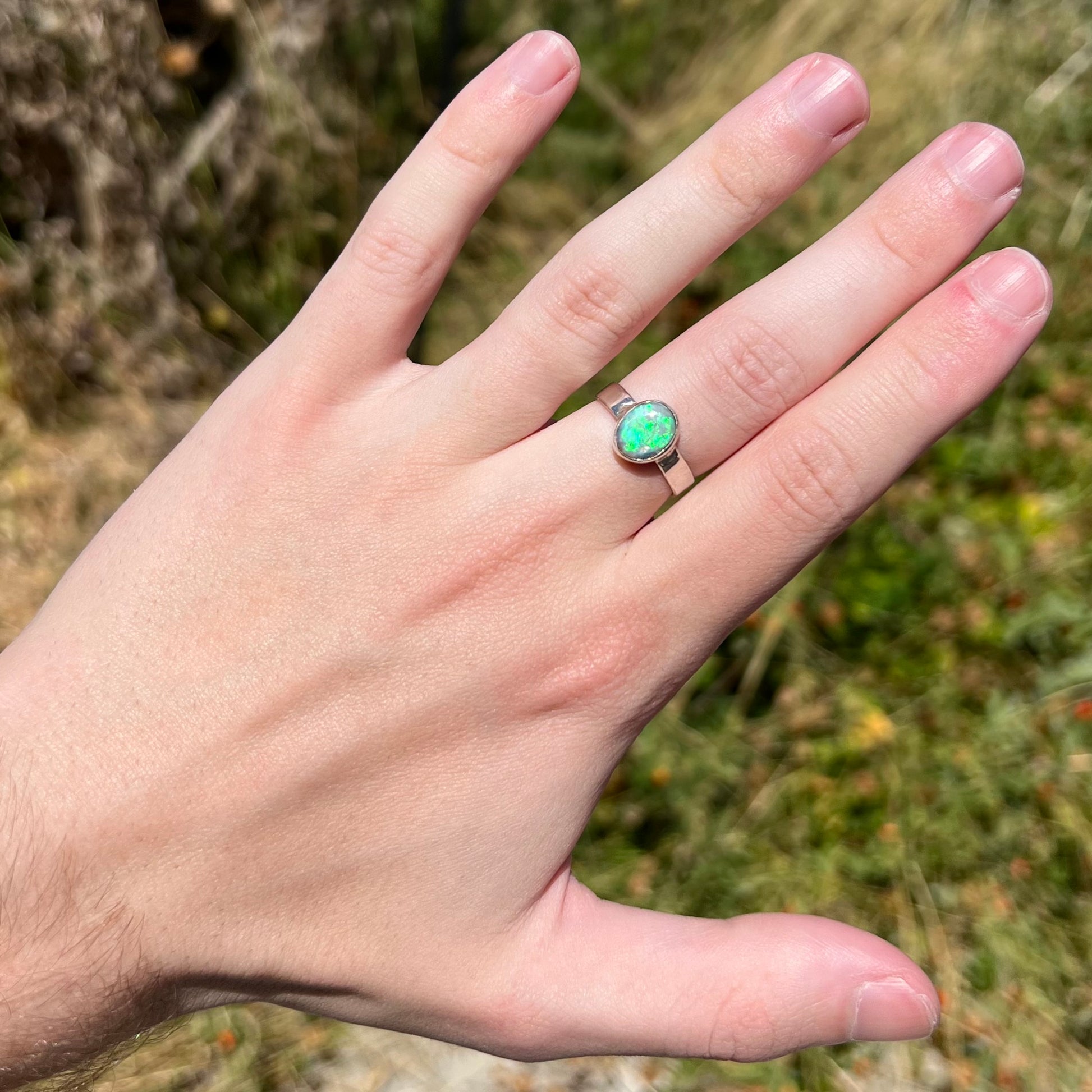 Hand wearing a natural Australian opal solitaire ring in sterling silver in outdoor sunlight; the opal has a green flash of color.