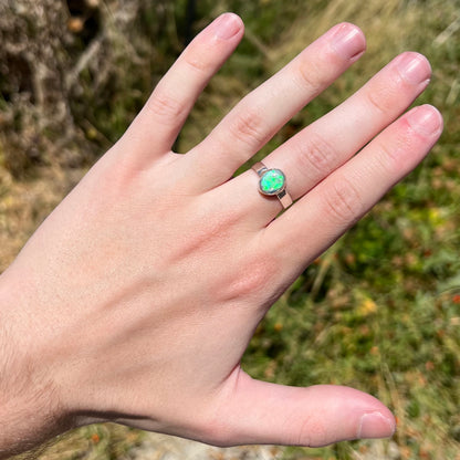 Hand wearing a natural Australian opal solitaire ring in sterling silver in outdoor sunlight; the opal has a green flash of color.