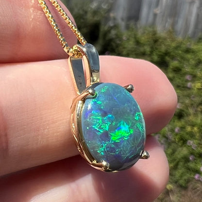 Angled sunlit close-up of natural Australian black opal pendant in 14k gold with green and blue flashes of color, resting on fingers