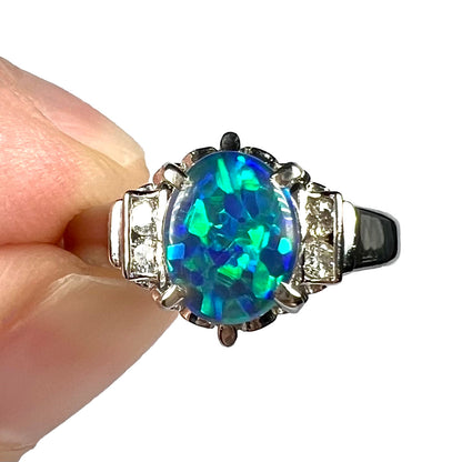 Natural Australian black opal and diamond platinum ring photographed face-up against a white background, held between two fingers.