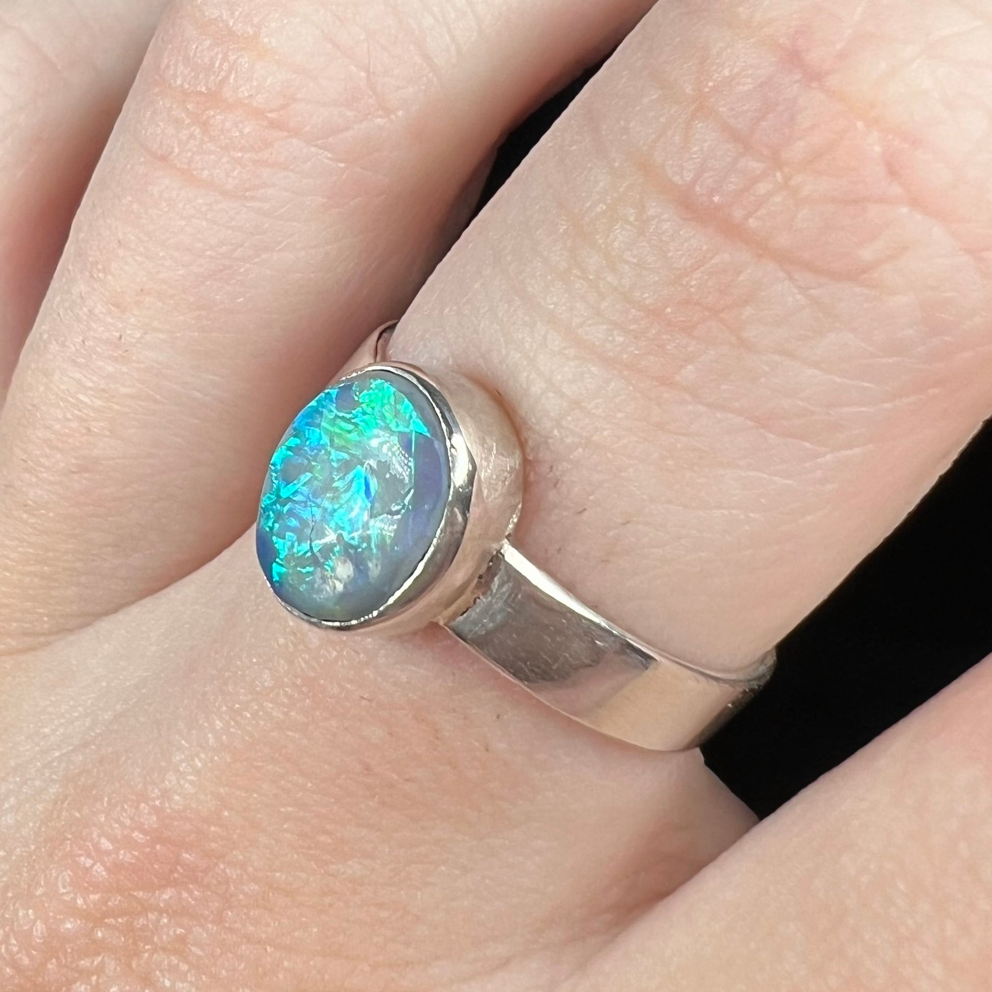 Angled close-up of a men's natural opal ring in sterling silver, worn on a finger; the opal has a bright blue flash of color.