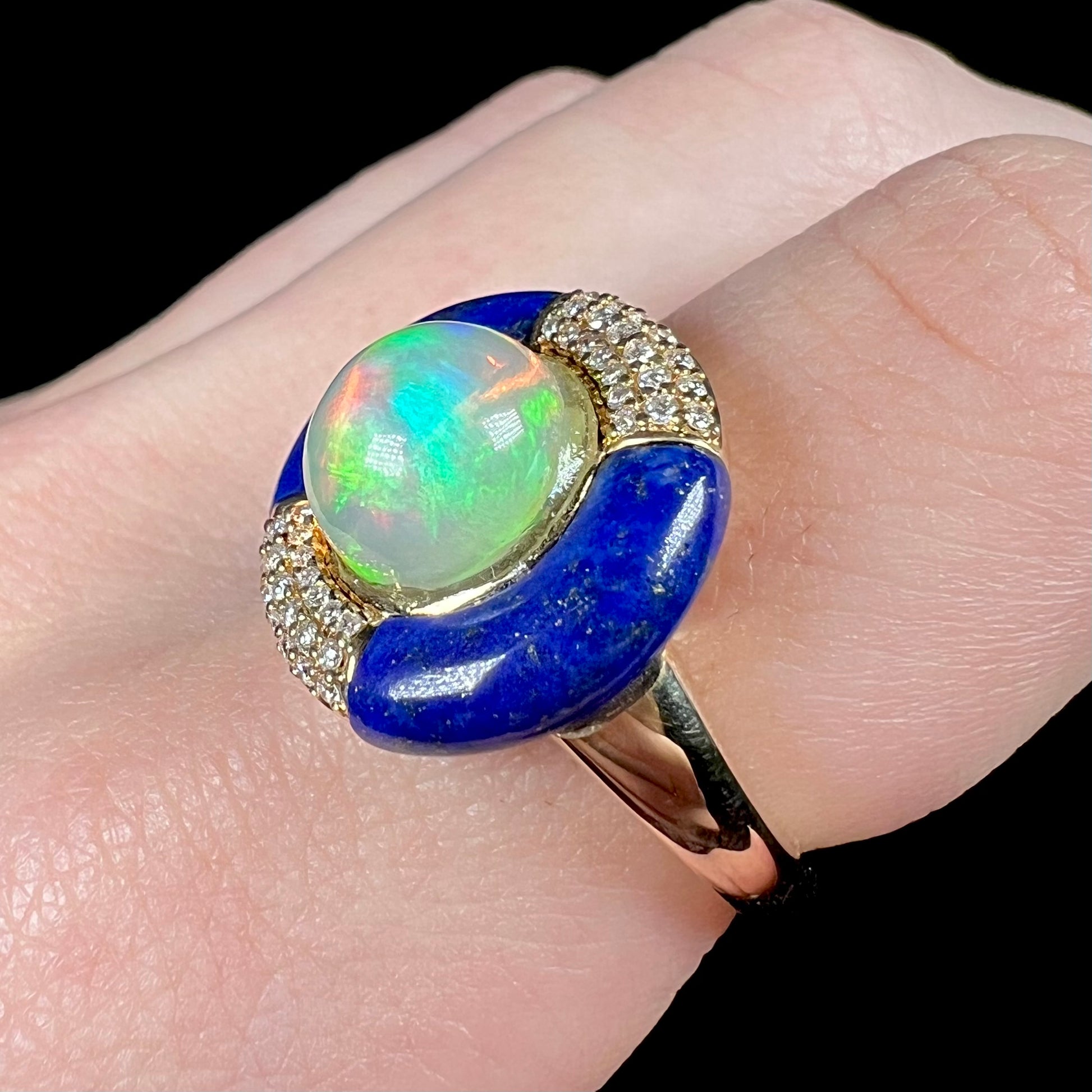 Angled close-up of a natural fire opal ring with a carved lapis lazuli and diamond pave halo in 14k yellow gold, worn on a finger.