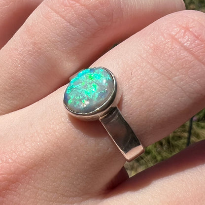 Angled close-up of a natural Lightning Ridge opal ring in sterling silver, worn on a finger in sunlight shining blue flashes of color.