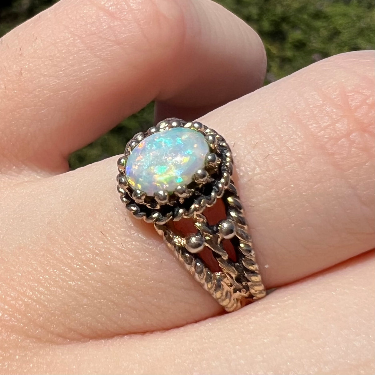 Angled close-up of a base metal costume solitaire ring set with a natural Australian opal, worn on a finger; the opal shows blue flashes of color in sunlight