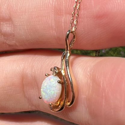 Side close-up of natural Australian opal and diamond pendant in dainty 14k gold asymmetrical design, held in sunlight