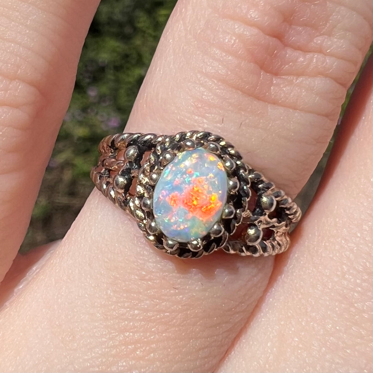 Cloe-up sunlit view of a natural Australian opal ring in base metal, worn on a finger; the opal has bright red and orange flashes of color