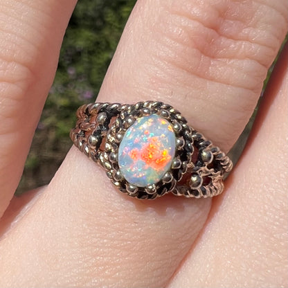 Cloe-up sunlit view of a natural Australian opal ring in base metal, worn on a finger; the opal has bright red and orange flashes of color