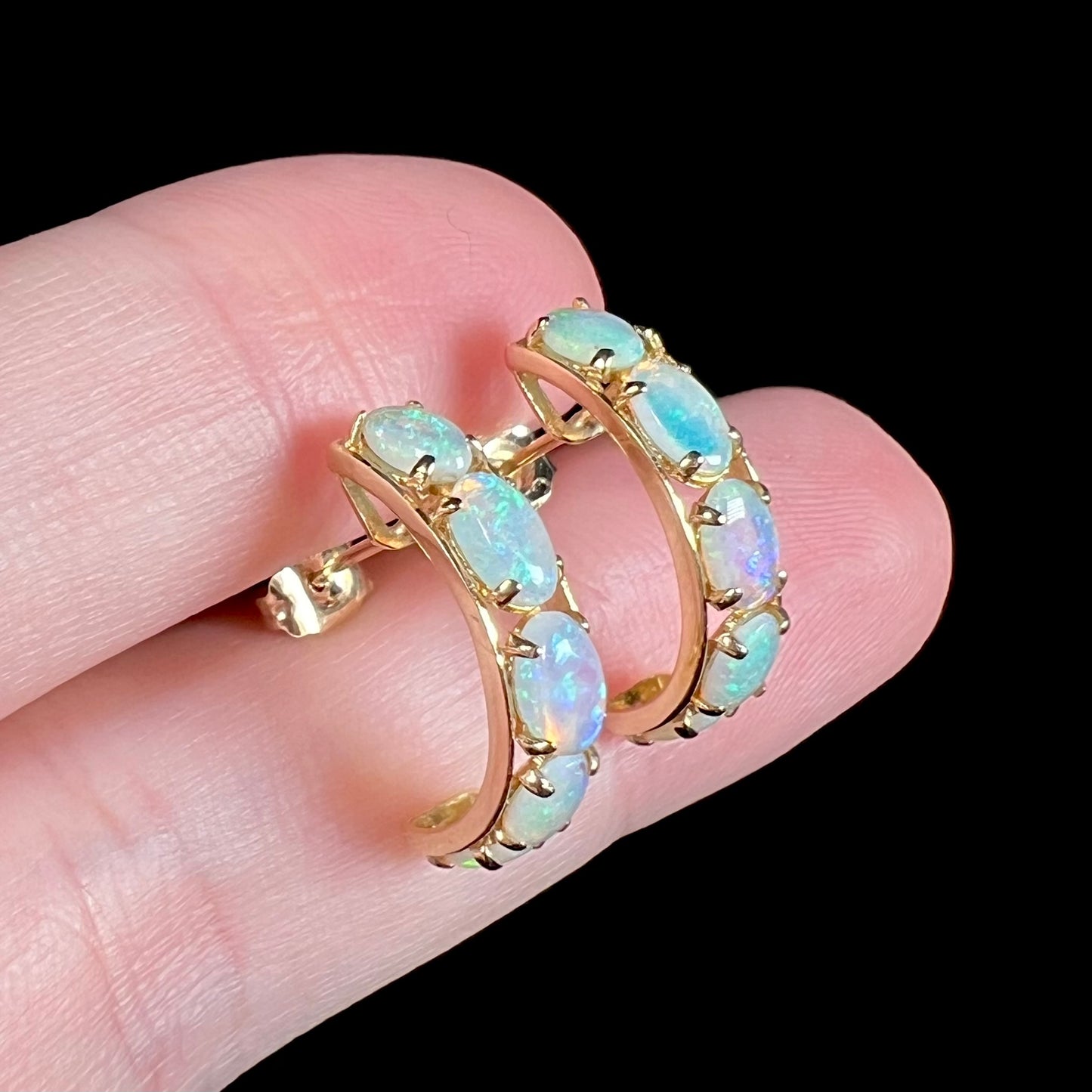 Angled close-up of natural opal semi-hoop earrings in yellow gold shown held between two fingers.  The opals have a bluish tint.