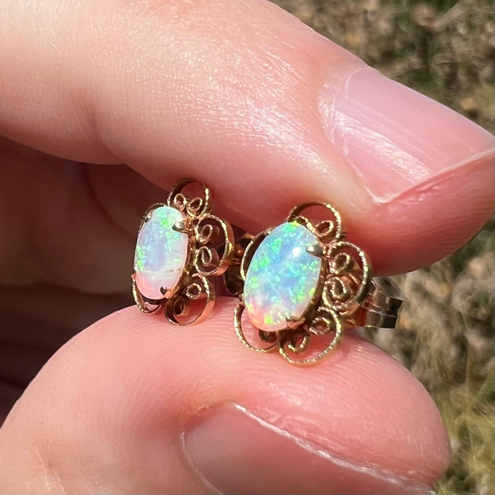 Pair of natural opal stud earrings with flower design in 14k yellow gold, shown from angle in sunlight between fingers