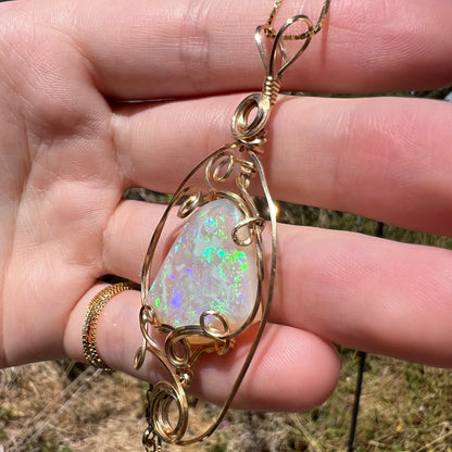 A gold wire-wrapped pendant necklace featuring a natural opal gemstone with iridescent green and blue flashes, shown from a close-up angle in a person's hand against an outdoor background in sunlight.