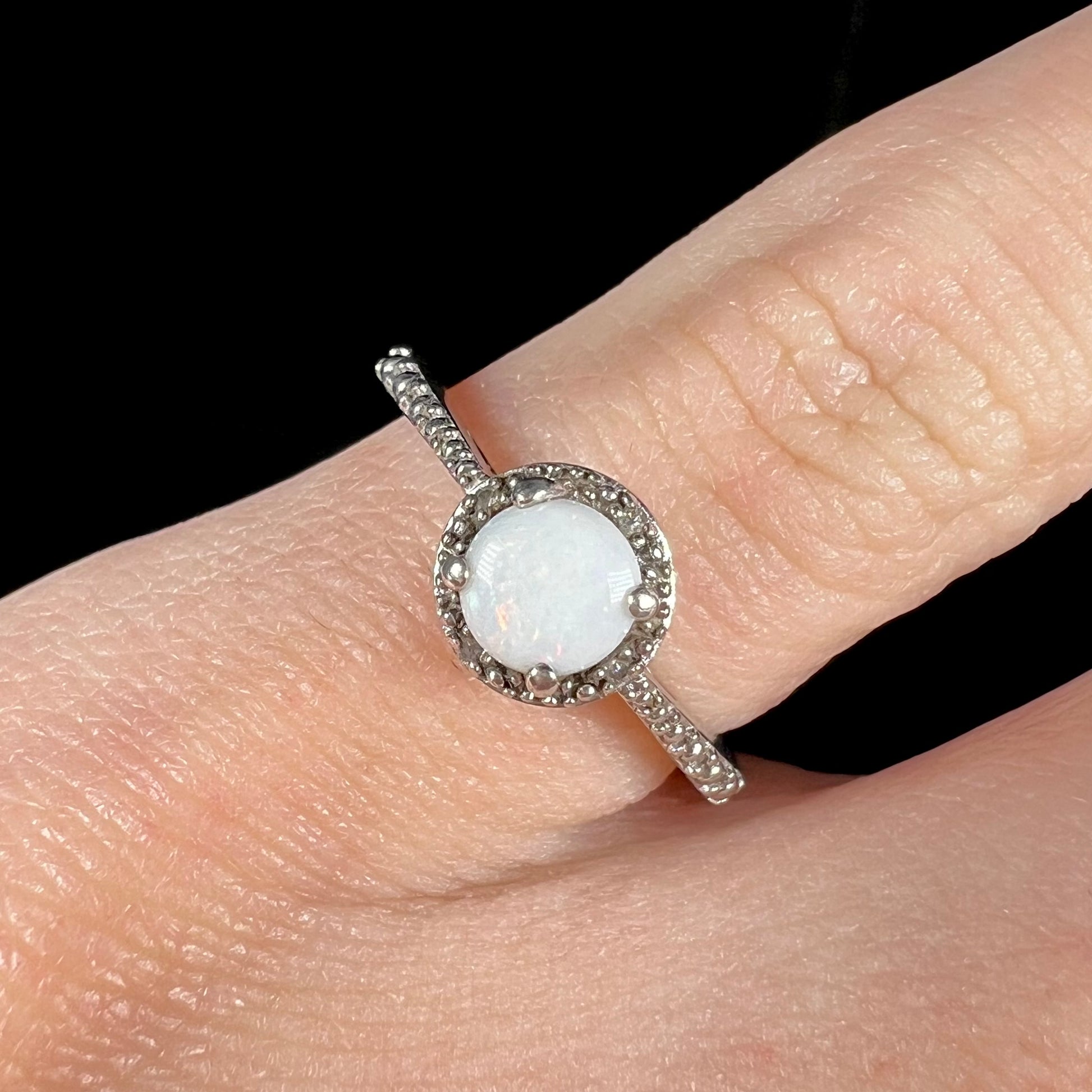 Close-up of a sterling silver white opal and diamond ring with a halo design, worn on a pinky finger.
