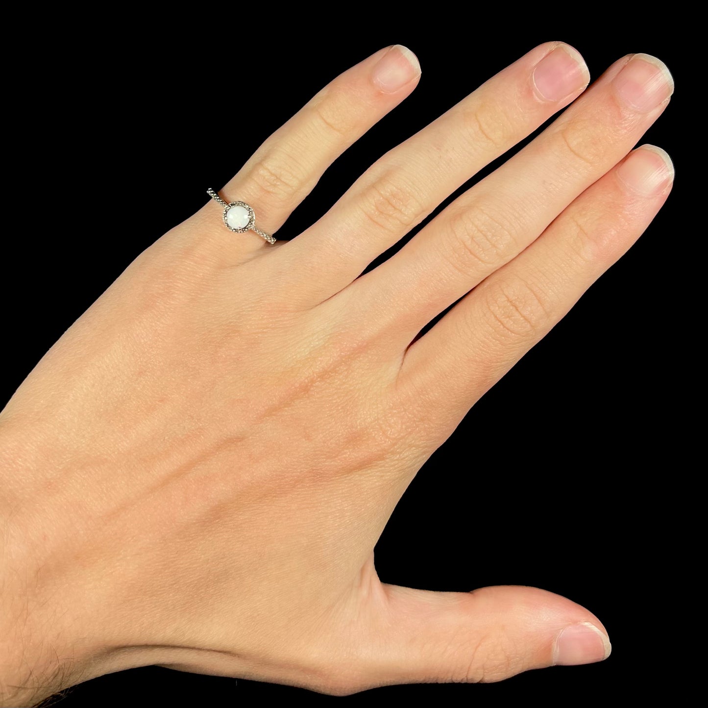 A sterling silver white opal and diamond ring with a halo design worn on a the pinky finger of a hand.