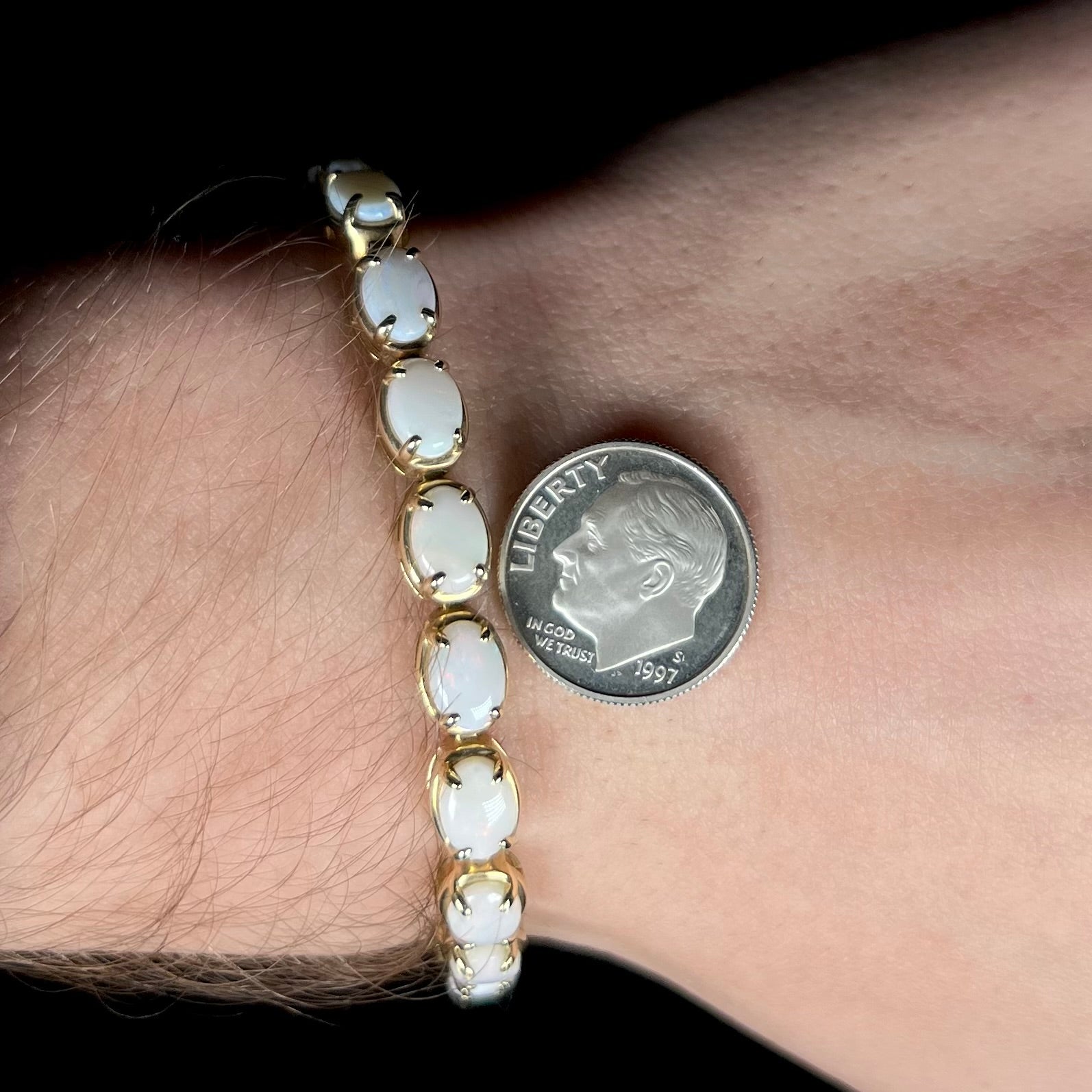 A natural yellow gold and white opal tennis bracelet worn on a wrist next to a US dime coin for scale.