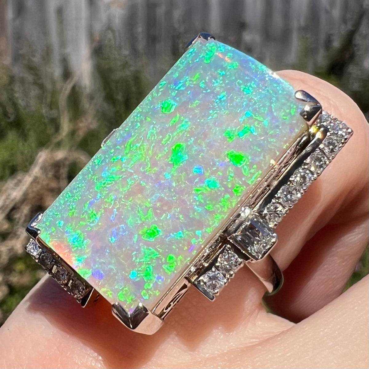 Angled close-up of 11.46ct rectangular opal and diamond cocktail ring worn on finger.  Sunlight shows the opal's non-directional green and blue play-of-color evenly distributed across the stone.
