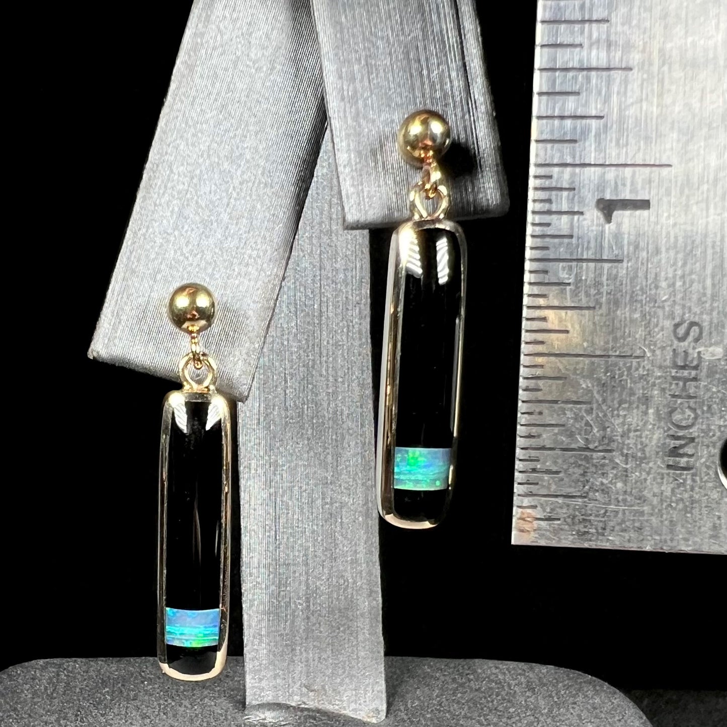A pair of gold drop earrings set with onyx and opal inlay that measure 1.25 inches in length, shown next to a ruler.