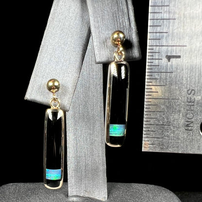A pair of gold drop earrings set with onyx and opal inlay that measure 1.25 inches in length, shown next to a ruler.