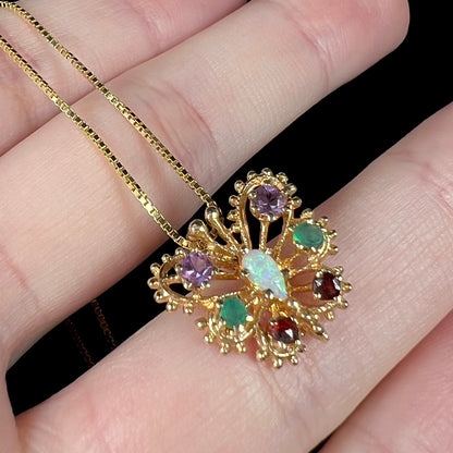 Angled close-up of an opal, amethyst, emerald, and garnet multi-gemstone butterfly pendant on gold tone chain in hand.