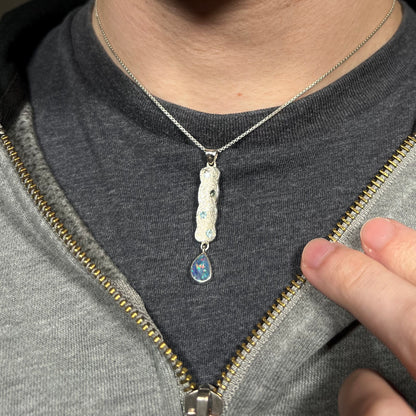 A sterling silver opal necklace with a blue topaz-accented dangling drop design, worn around a person's neck against a dark shirt.