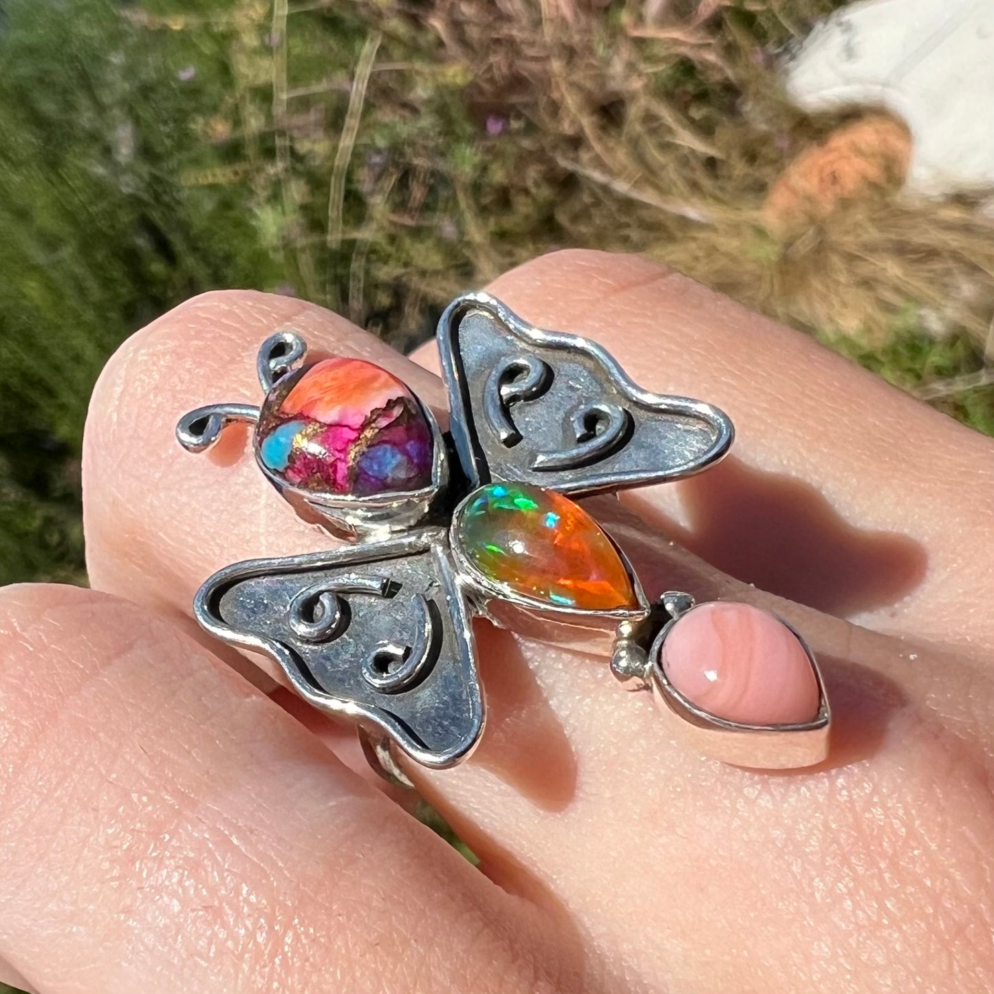 An opal butterfly ring made from silver, worn on a hand and shown at a close-up angle in sunlight.