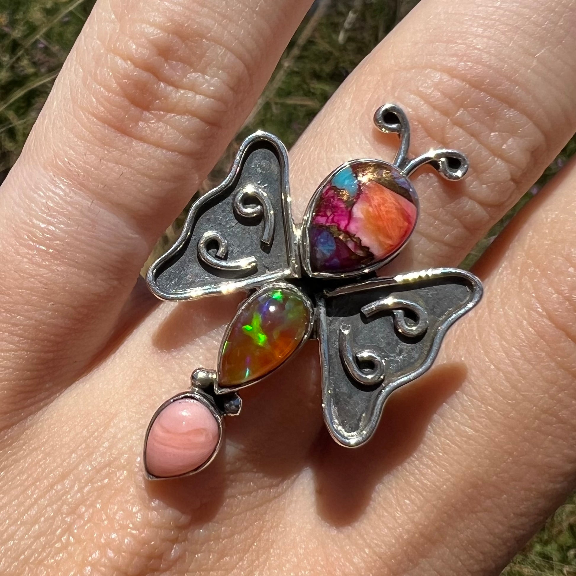 Close-up of an opal butterfly ring, worn on a hand in the sunlight and shown from the front.