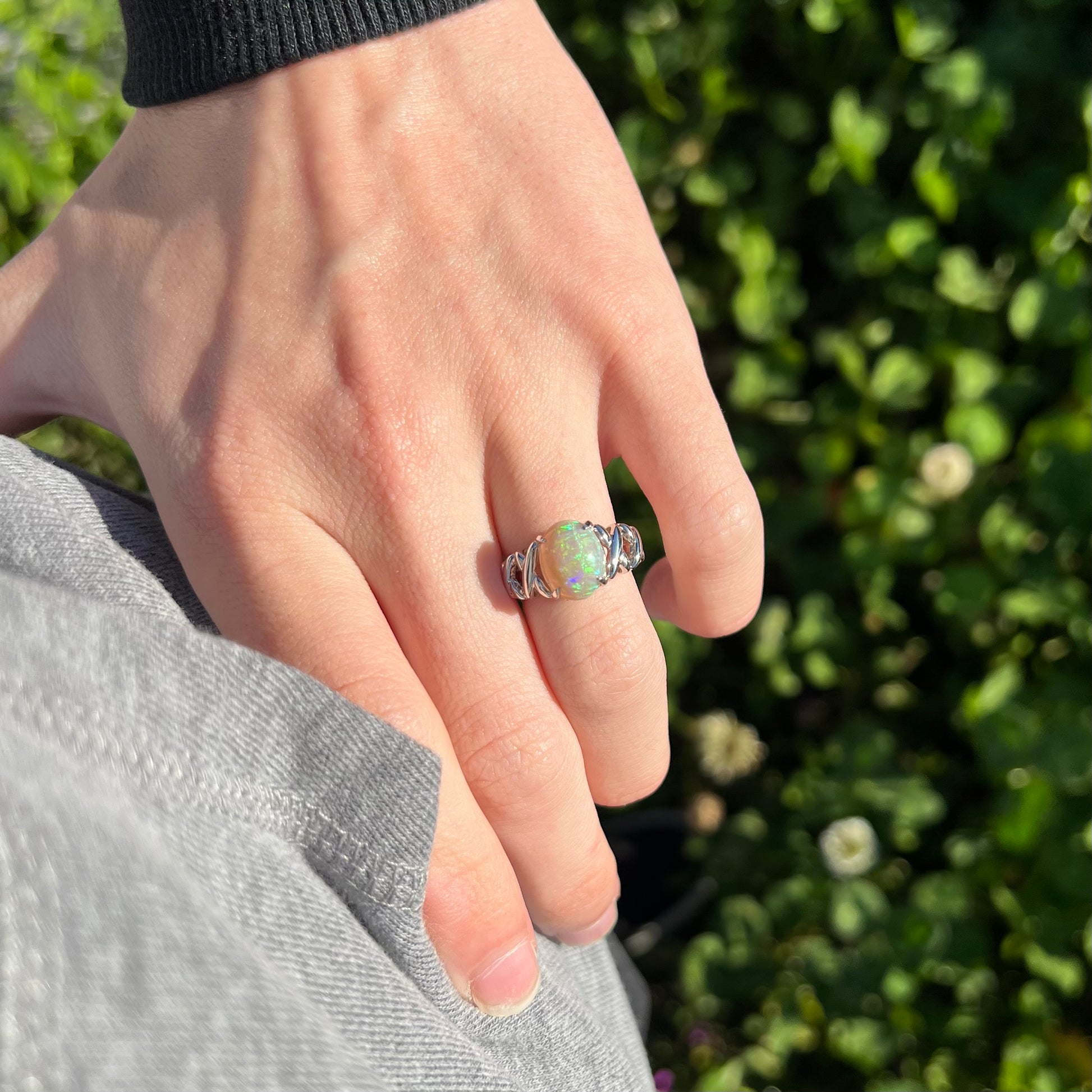 A platinum opal crisscross ring worn on a hand against a model's hip in the sunlight.