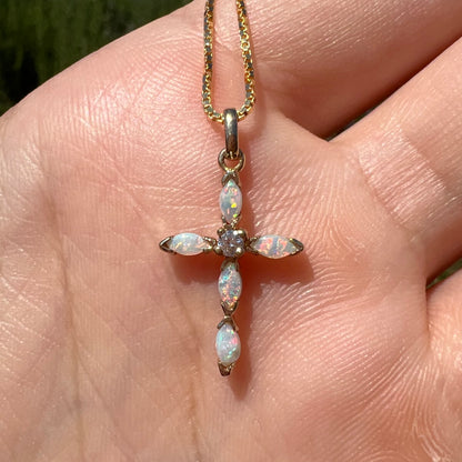 A dainty opal cross necklace shown in a hand, straight on, close-up in the sunlight.