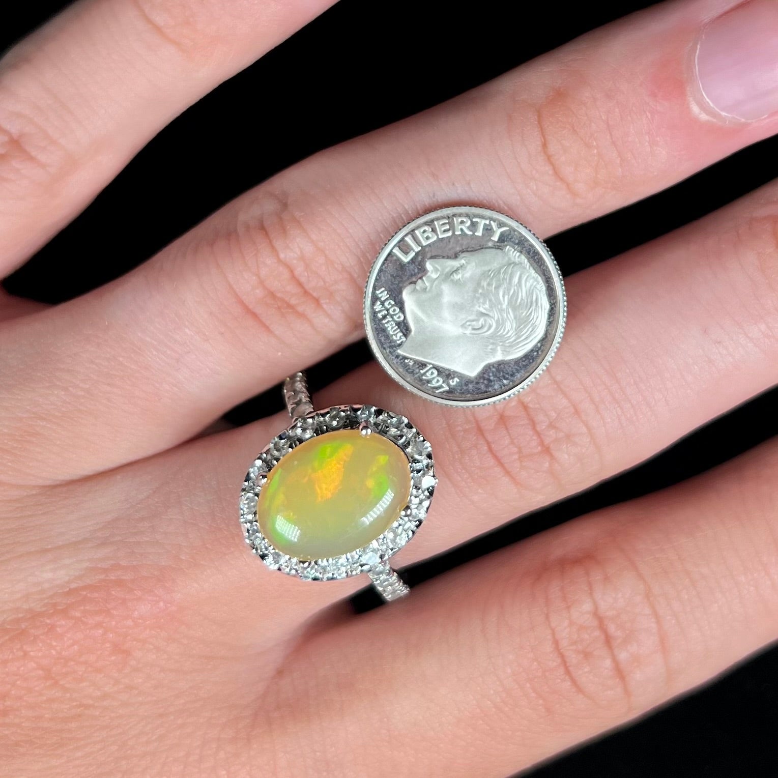 A sterling silver Ethiopian fire opal ring with a CZ halo worn on a hand next to a 1997 S dime coin.