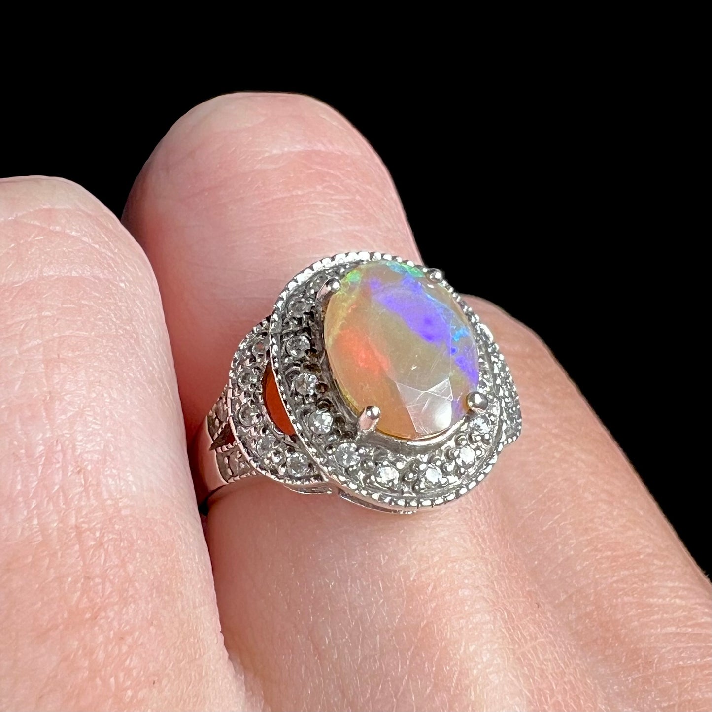 An Ethiopian opal and CZ halo ring worn on a finger, shown close-up at an angle.  The opal shows purple color play.