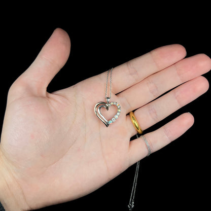 A two-tone sterling silver and yellow gold lab-created opal and CZ heart necklace, held in a hand against black.