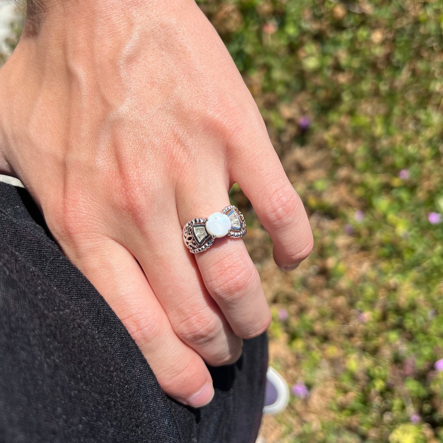 A round Australian white opal and CZ ring in white base metal worn on a hand resting on the model's hip.