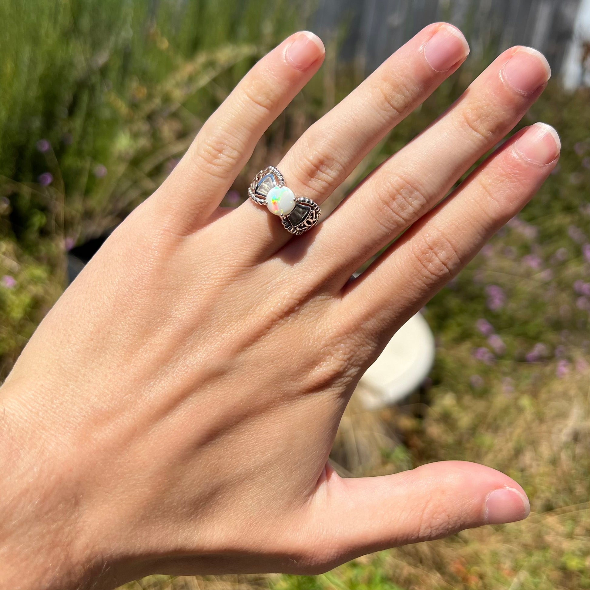 A round Australian white opal and CZ ring in white base metal worn on a hand in the sunlight.