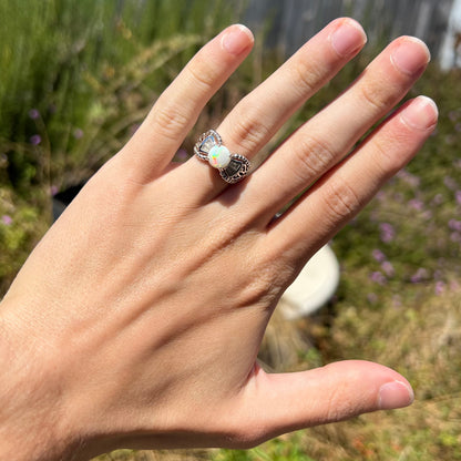 A round Australian white opal and CZ ring in white base metal worn on a hand in the sunlight.