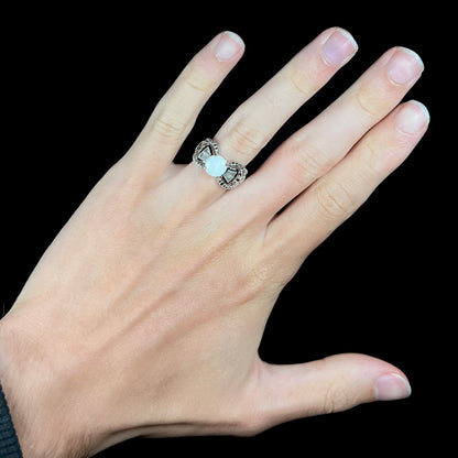 A round Australian white opal and CZ ring in white base metal worn on a hand against black background.