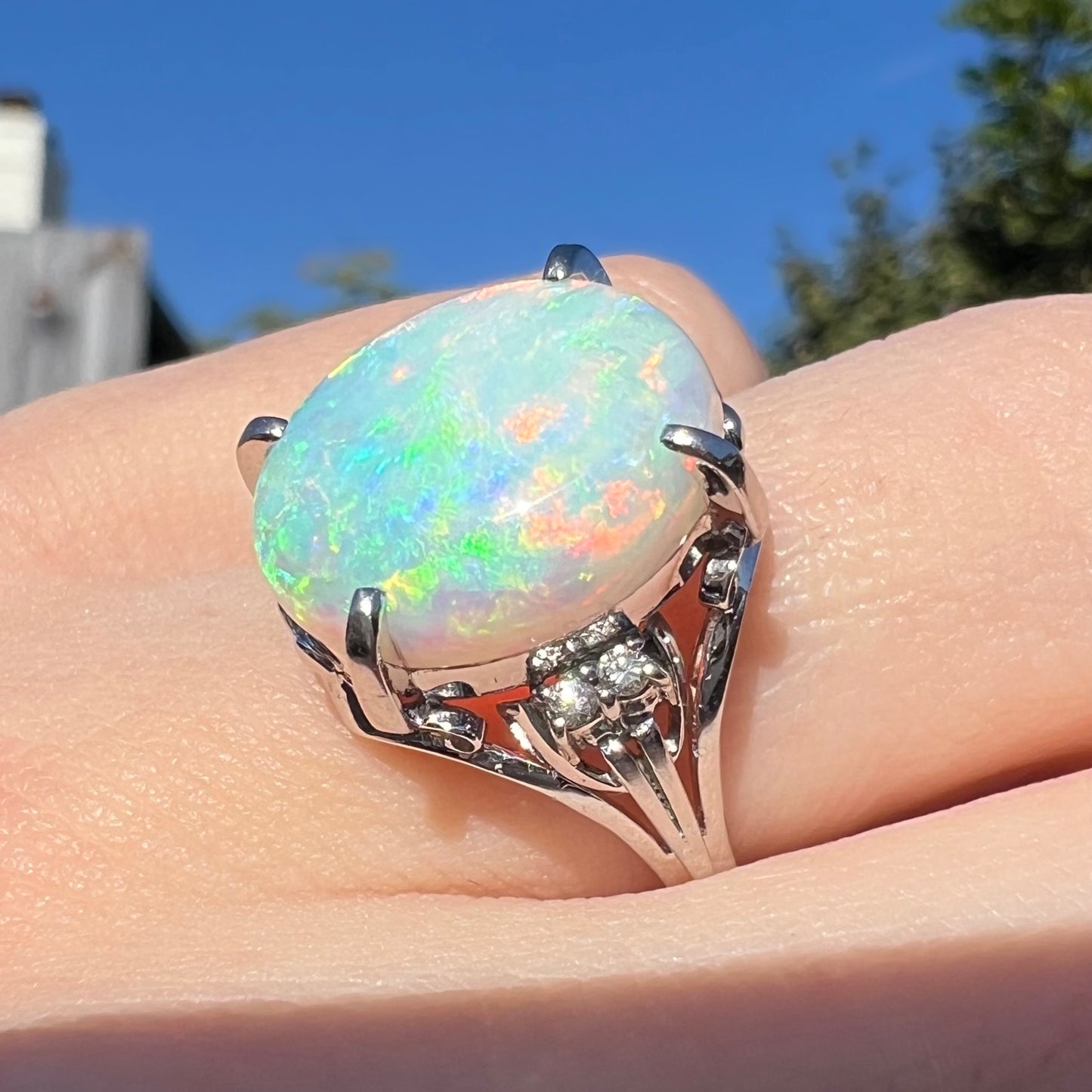 A large opal and diamond engagement ring worn on a finger in sunlight.