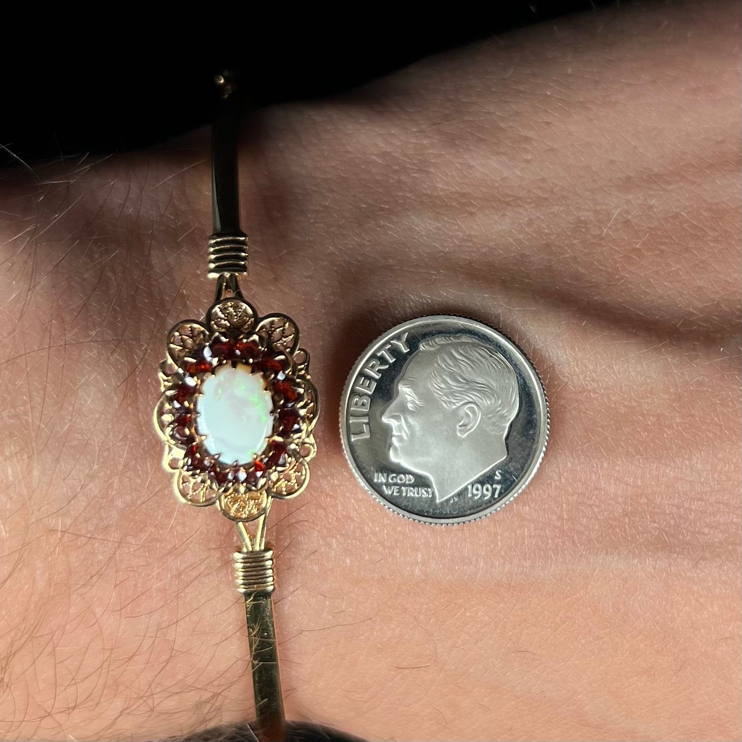 A vintage yellow gold Australian opal and garnet halo bangle bracelet shown close-up on a wrist next to a dime coin.