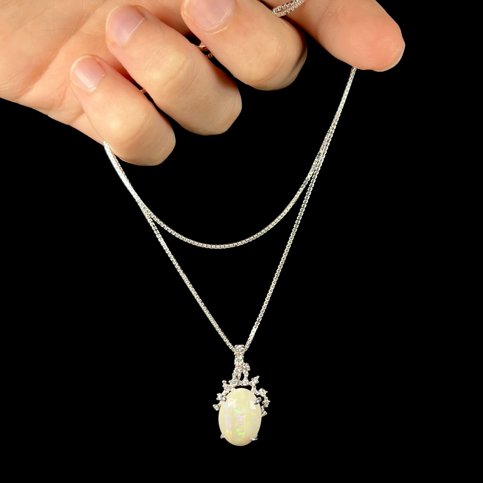 An opal and diamond necklace being dangled by the chain in a hand against a black background.