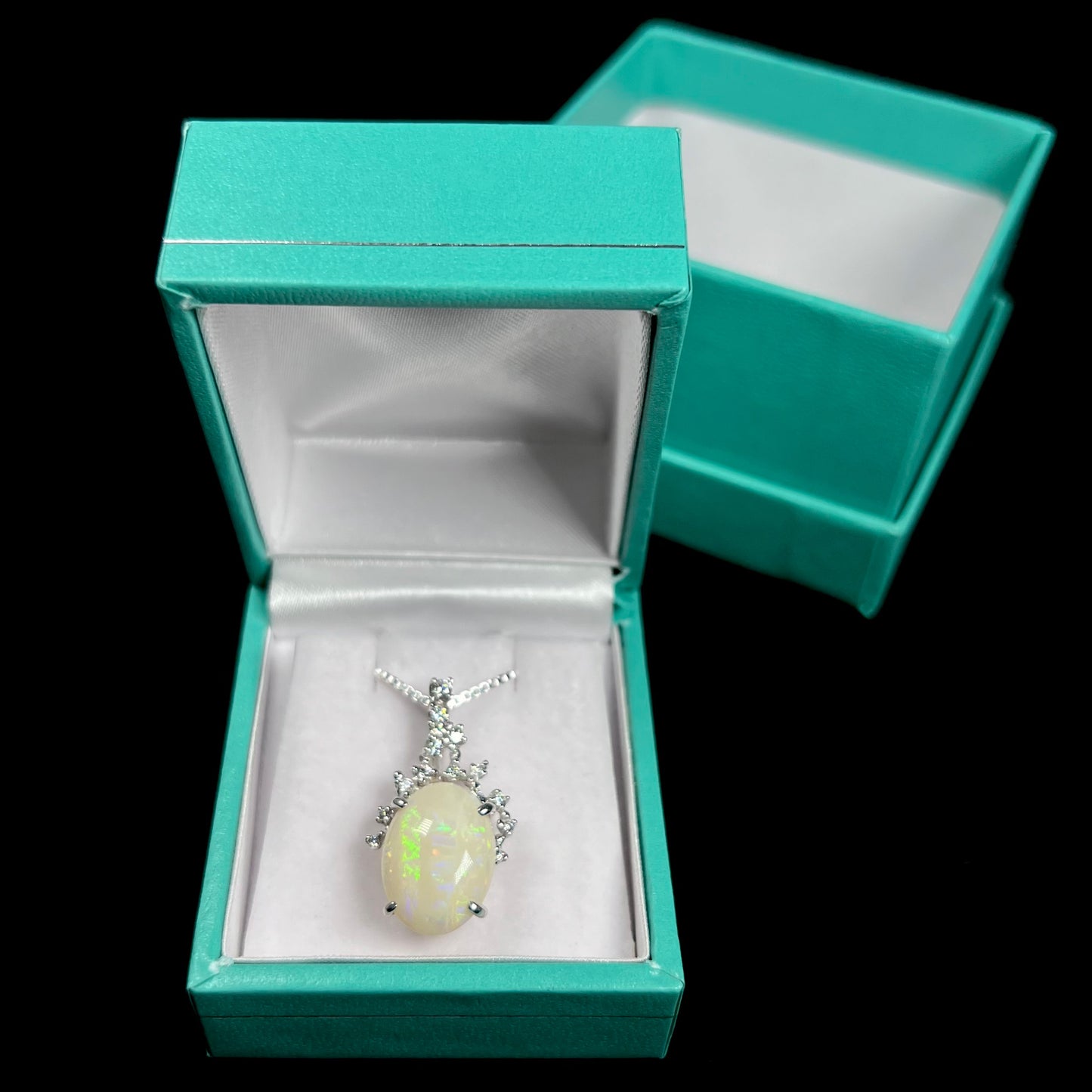 An 18k white gold opal and diamond necklace displayed inside a blue pendant box, with a vibrant green stripe visible in the opal.