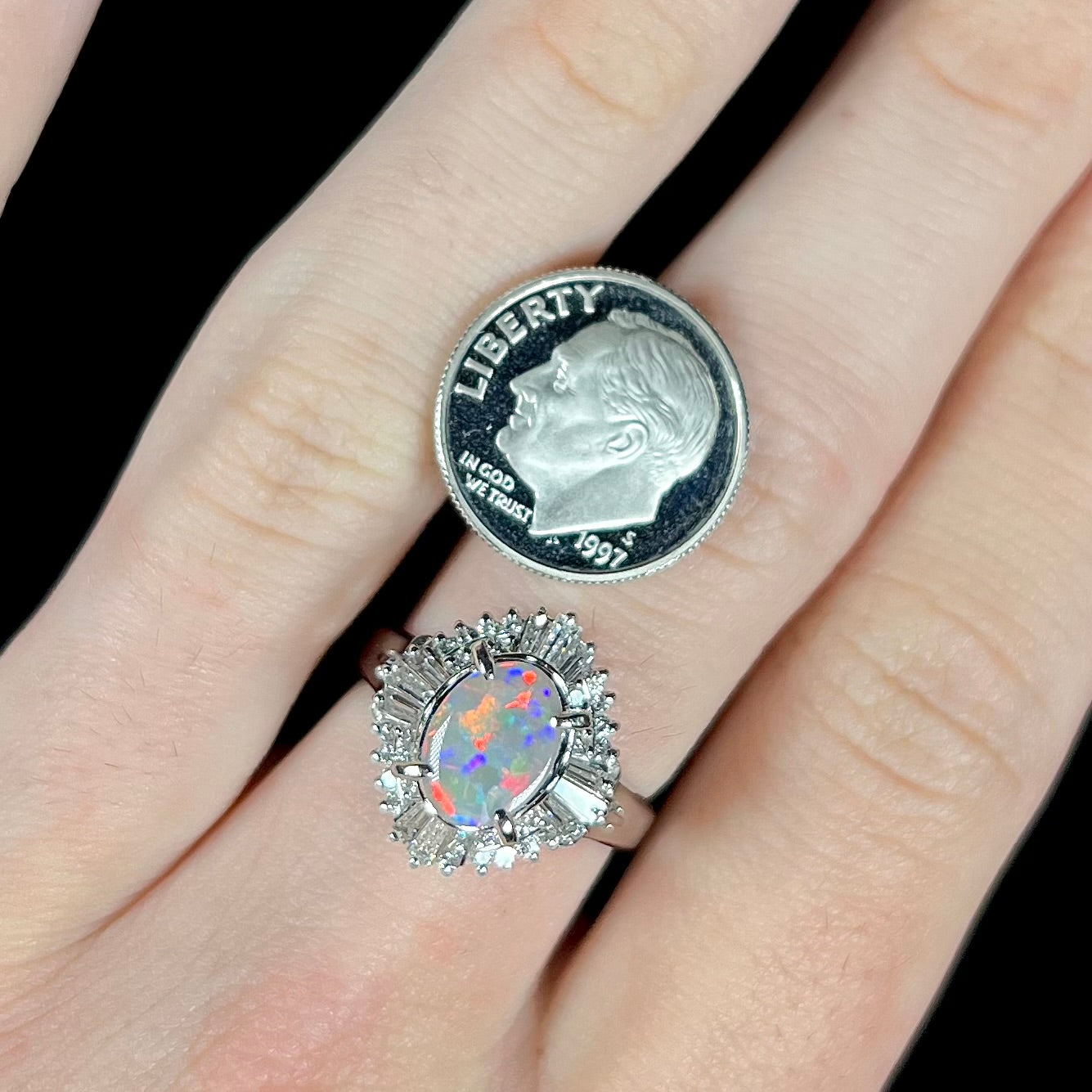 A semi-black opal and diamond spray-design ring in platinum, worn on a hand next to a US dime coin for scale.