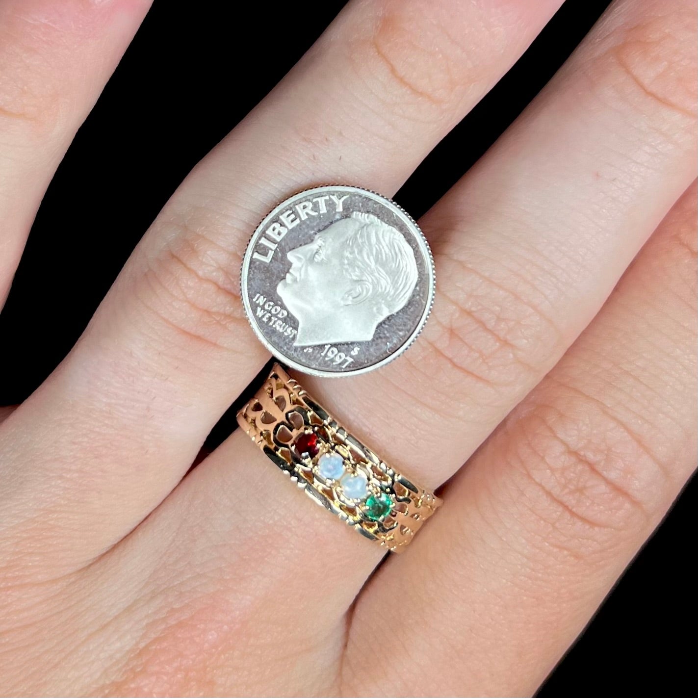 A gold woven-style mother's ring set with two opals, an emerald, and a garnet worn on a hand next to a dime coin.