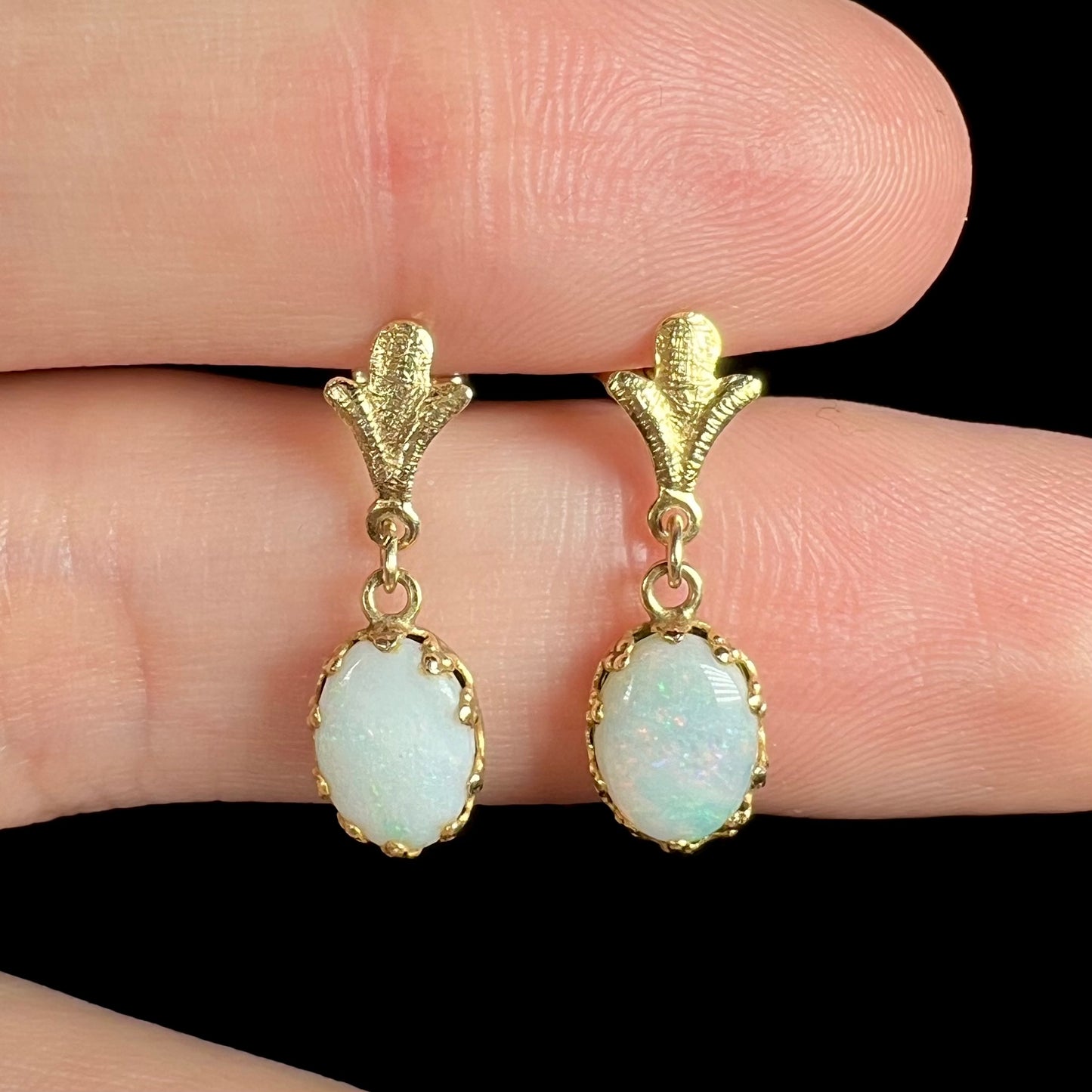 Close-up of a pair of yellow gold Fleur-de-Lis Australian light opal dangle earrings, shown held between two fingers.