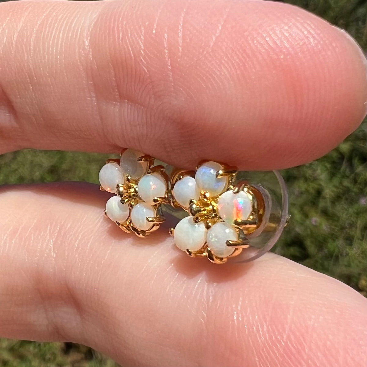 Australian opal flower earrings in gold-tone metal shown close-up in sunlight between two fingers