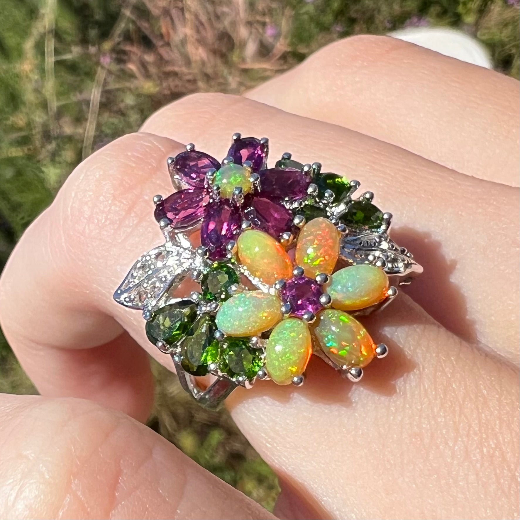 An opal flower ring worn on a finger at an angle outdoors in natural light, shown close-up.